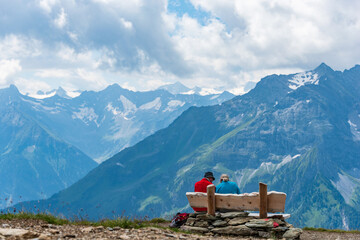 Two older people, tourists, rest on a bench in the mountains. Around them there is a view of the majestic snow-covered Alps. - 403635110
