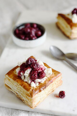 Puff pastry squares with ricotta and cherry sauce on white marble tray. Baked small square cakes topped with cheese and berry sauce