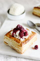 Puff pastry squares with ricotta and cherry sauce on white marble tray. Baked small square cakes topped with cheese and berry sauce