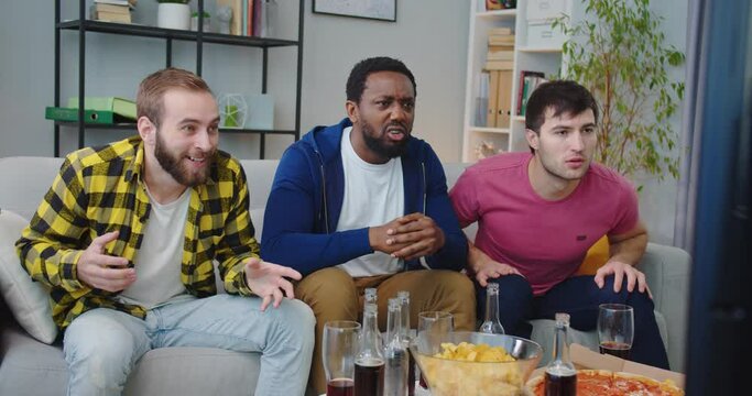 Young happy emotional African American and Caucasian guys friends together watching match on TV at home. Men screaming, hugging and celebrating victory cheering fav team. football fans, scoring goal
