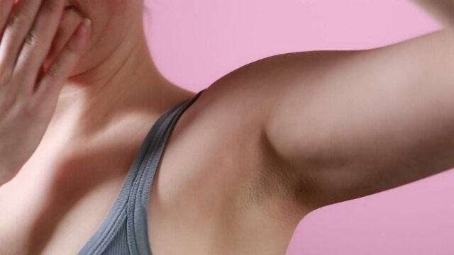 Hygiene and body care. The woman leans down to sniff her armpit and turns away sharply because of the bad smell. Close-up of the armpit. Pink background