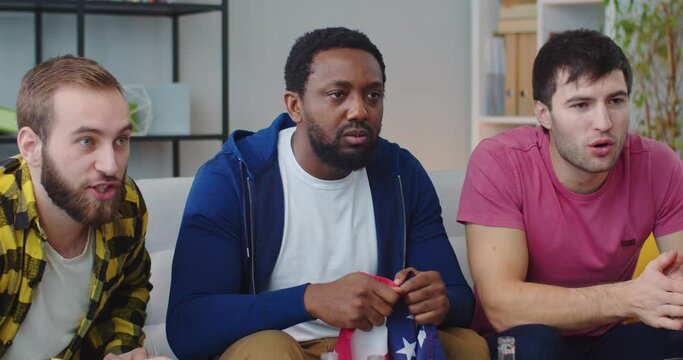 Young cheerful emotional mixed-races guys friends together watching soccer match on TV at home holding USA flag. Men screaming and celebrating victory cheering fav team. football fans, scoring goal