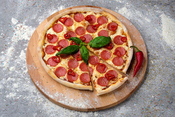 Italian pepperoni pizza served with fresh basil leaf and spicy whole chili pepper on wooden plate decorated with culinary flour. Fast food dish with salami slices on concrete stone background
