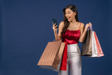 Young Asian woman using mobile phone for online shopping on blue color background, black friday, cashless payment and consumerism concept, carrying shopping bags