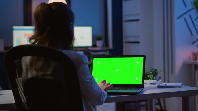 Back view of business woman looking at green screen laptop sitting at desk in start up office overworking. Freelancer watching desktop monitor display with green mockup, chroma key, during night time