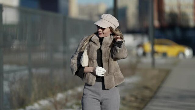 A young smiling blonde woman in sunglasses, in a sheepskin coat walks and poses on a sunny city street. Winter fashion.