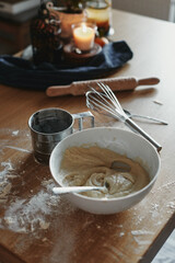 White bowl with dough and spoon in it, on the wooden table in the kitchen. The process of mixing ingredients for pastry. Closeup, selective focus.