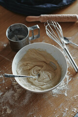 White bowl with dough and spoon in it, on the wooden table in the kitchen. The process of mixing ingredients for pastry. Closeup, selective focus.