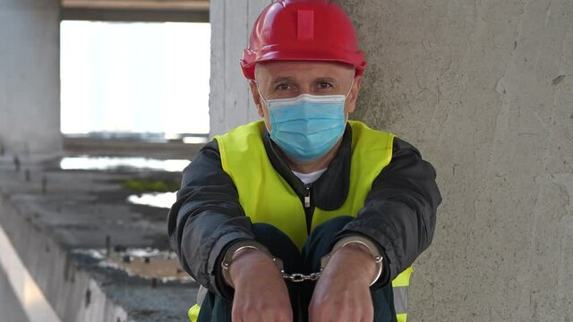Handcuffed worker in medical protective mask and red hard hat sits at construction site and looks at the camera