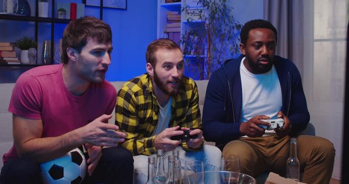 Cheerful happy African American and Caucasian men playing video games football sitting on couch laughing together. Leisure concept. Guys best friends play video game on console at night having fun