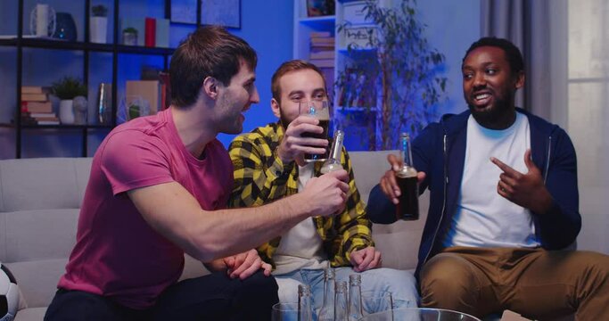 Portrait of mixed-races joyful happy male best friends spending evening together at home sitting on couch with beer drinks having fun chatting and laughing in good mood. Leisure concept