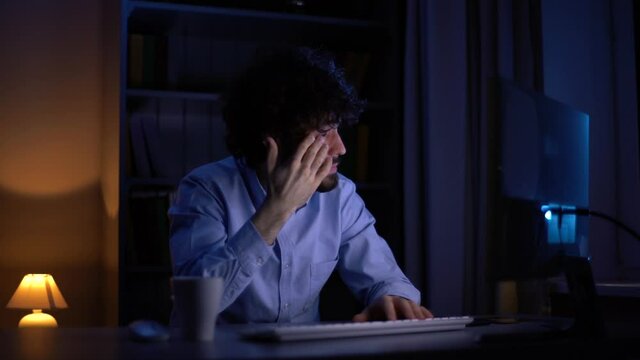 Sleepy young bearded man working late at night on computer rubbing eyes at home office in dark room. Tired sleepy student prepares for late night session. Young freelancer working late over project.