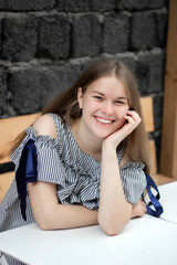 Smiling young woman in blue dress sitting on wooden bench and leaning on white table in front of black brick wall, looking at camera. Girl having fun. Urban style and fashion. Summer casual outfit.