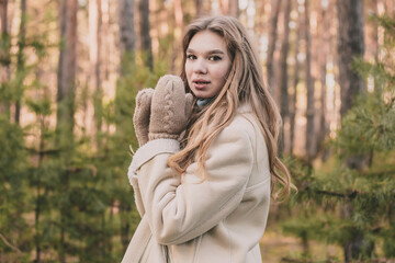 a young girl of model appearance in a pine forest is dressed in a fur coat and a warm sweater shows hands in mittens and smiles