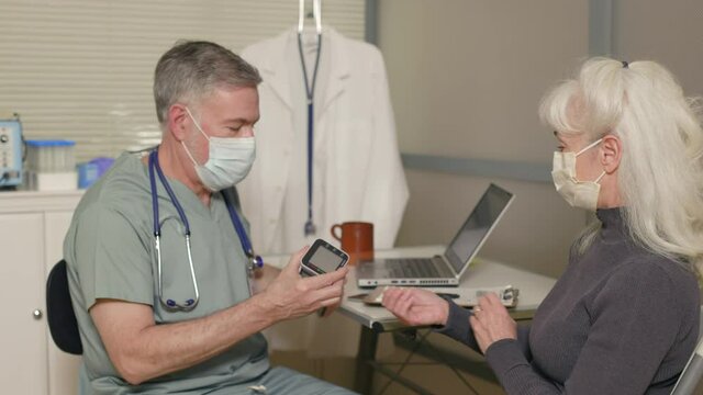 A doctor and patient both wearing masks due to COVID-19 sitting in a clinical setting involved in a procedure using a small device that measures blood pressure.