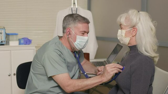 A doctor and patient both wearing masks due to COVID-19 involved in a procedure using a stethoscope to listen to condition of heart and lungs capacity then documenting the results.