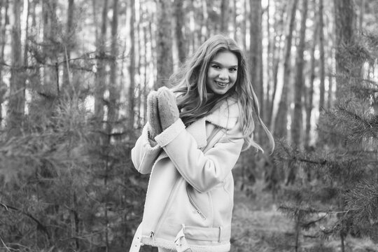 
a young girl of model appearance in a pine forest is dressed in a fur coat and a warm sweater holds her hands in mittens black and white photo