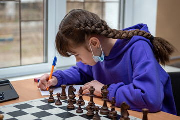 Teenage girl in a medical mask at the chessboard. Game of chess. Figures on a chessboard. Chess tournament during a pandemic.