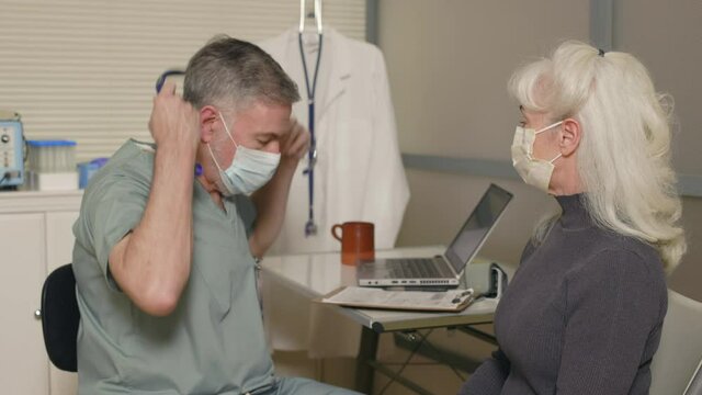 A doctor and patient both wearing masks due to COVID-19 sitting in an exam room involved in a procedure using a stethoscope to listen to condition of heart and lungs capacity.
