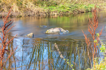 Close-up of water stream flowing between stones in a river between grass and wild plants, autumn day in Sweikhuizen, South Limburg, the Netherlands. Long exposure photography