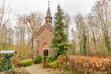 Naklejka premium Chapel of St. Anna (St. Annakapel) with its brick walls and a small tower among bushes, a green pine and bare trees in the background, cloudy winter day in Spaubeek, South Limburg, the Netherlands