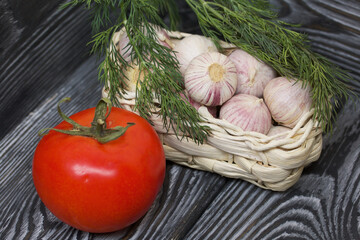 Garlic in an overturned wicker basket. Dill sprigs and tomato. On black pine boards. Close-up shot.