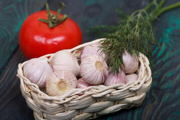 Garlic in an overturned wicker basket. Dill sprigs and tomato. On green pine boards. Close-up shot.