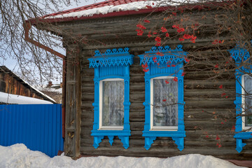 windows of a wooden house with blue carved platbands on a winter day. rowan with red berries grows under the windows. Cozy home