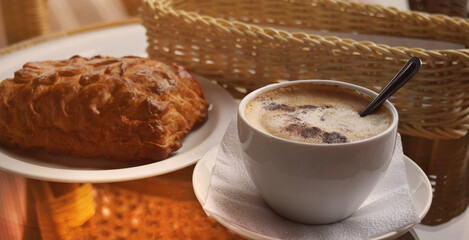 A cup of coffee with a fresh bun on a glass table in a cafe in the early morning. Selective focus. Soft light. Comfort.
