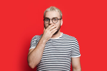 Blonde man is surprised by something covering his mouth and looking at camera on a red studio wall wearing glasses - obrazy, fototapety, plakaty