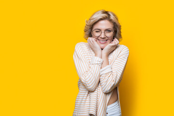 Blonde woman with glasses is advertising something on a yellow studio wall with free space smiling and touching her chin