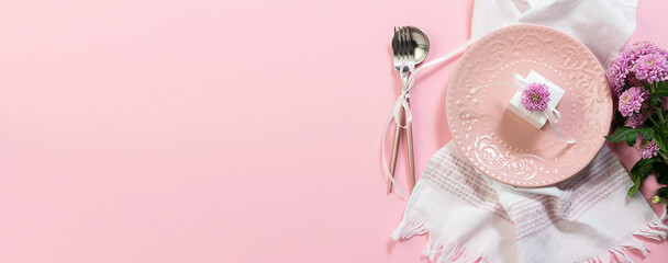 Festive table setting concept with pink plate, little present box. cutlery set and flowers on pink pastel background top view. Copy space for your text. long banner format.