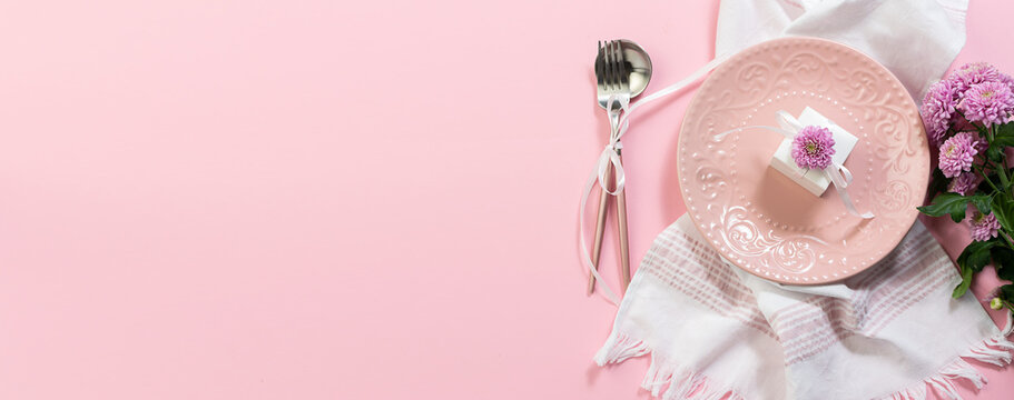 Festive Table Setting Concept With Pink Plate, Little Present Box. Cutlery Set And Flowers On Pink Pastel Background Top View. Copy Space For Your Text. Long Banner Format.