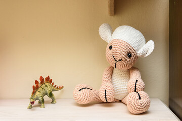 Ovelha sentada sobre uma estante de madeira ao lado um pequeno dinossauro. Ovelha feita na técnica de crochê amigurumi. © DR Moura