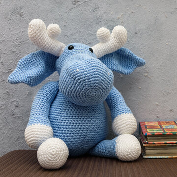 Alce azul de pel&uacute;cia, feito na t&eacute;cnica de croch&ecirc; amigurumi visto de frente sentado sobre um trilho marrom, fundo cinza.