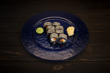 Top view of Japanese sushi roll served with ginger and wasabi on dark blue glass plate, wooden background. salmon, cucumber, cream cheese, flying fish roe wrapped in black rice and mamenori 
