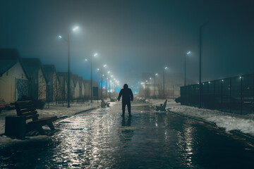 Fototapeta na wymiar Silhouette of dark man in hood in night illuminated city alley in foggy weather, misty horror and scary atmosphere concept.