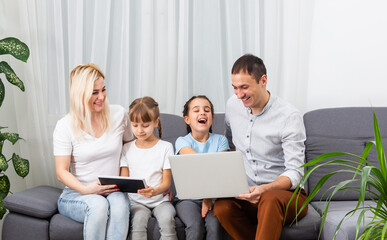Young joyful casual family of two kids and couple sitting on sofa and watching funny video or cartoons in touchpad