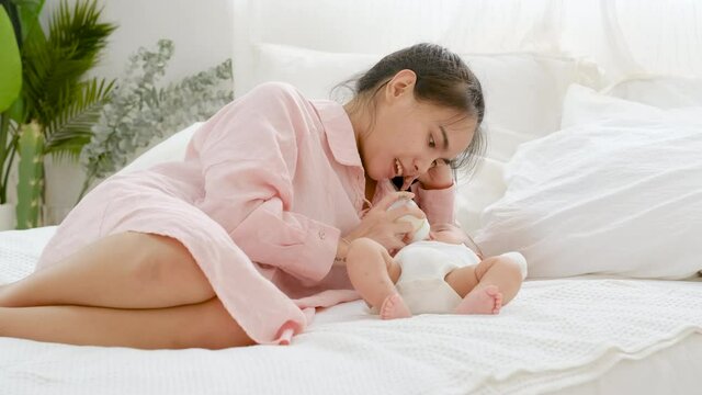Young Asian mother lie near her newborn baby to give milk via bottle to her baby and look carefully and love action of woman with healthcare and bonding in family concept.