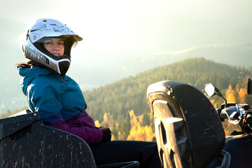 Happy woman driver in protective helmet enjoying extreme riding on ATV quad motorbike in summer mountains at sunset.