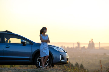 Happy young woman in blue dress standing near her vehicle looking at sunset view of summer nature. Travelling and vacation concept.