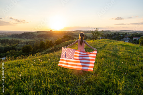 Back view of happy young woman posing with USA national flag standing outdoors at sunset. Positive girl celebrating United States independence day. International day of democracy concept.