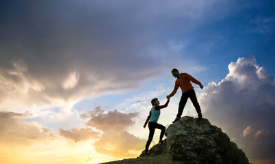 Man and woman hikers helping each other to climb a big stone at sunset in mountains. Couple...
