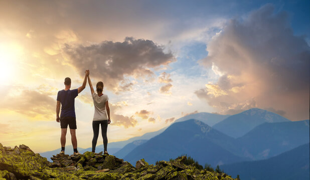 Back view of young hiker couple standing with raised arms holding hands on rocky mountain top enjoying sunset panorama.