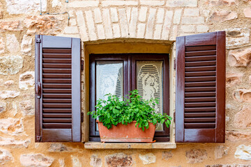 Small window of an old stone house, with brown shutters and pot with plant on the windowsill
