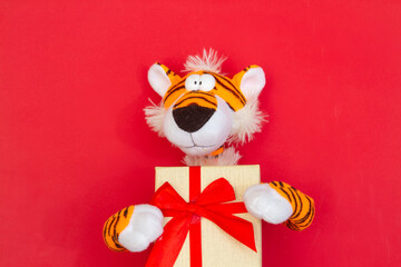 Year of the tiger 2022, toy tiger holding a gift with a red bow on a bright red background, holiday concept, soft cloth animal.

