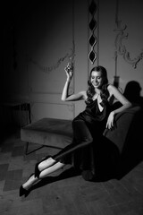Elegant lady in black long evening dress sitting at sofa in black silk dress and holding glass of sparkling wine. Celebrating concept.