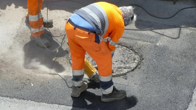 Teamworkers in protective orange uniform using drilling machine for destroying road asphalt for sewerage reconstruction, cement removal with powerful electric drill machine. Workmen using modern