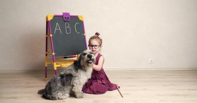 a little girl teacher plays with her dog and learns English letters with her . The girl sits next to the pet on the floor near the blackboard and praises it, strokes it and hugs it tightly