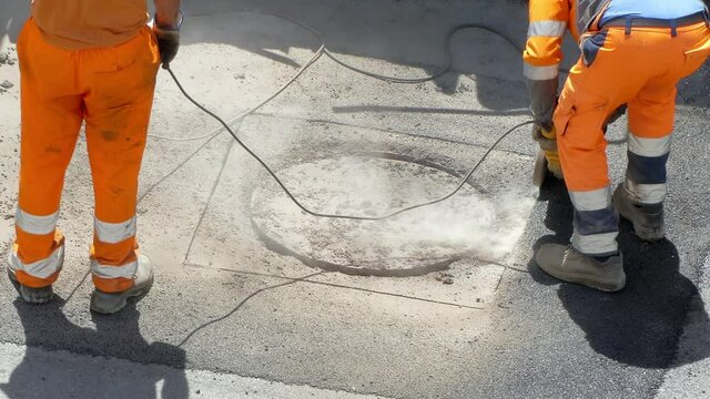Drilling works on the construction site, unrecognizable workers in orange uniform breaking asphalt with electric cutting machine causing dust pollution in the city. Men using drilling tools for